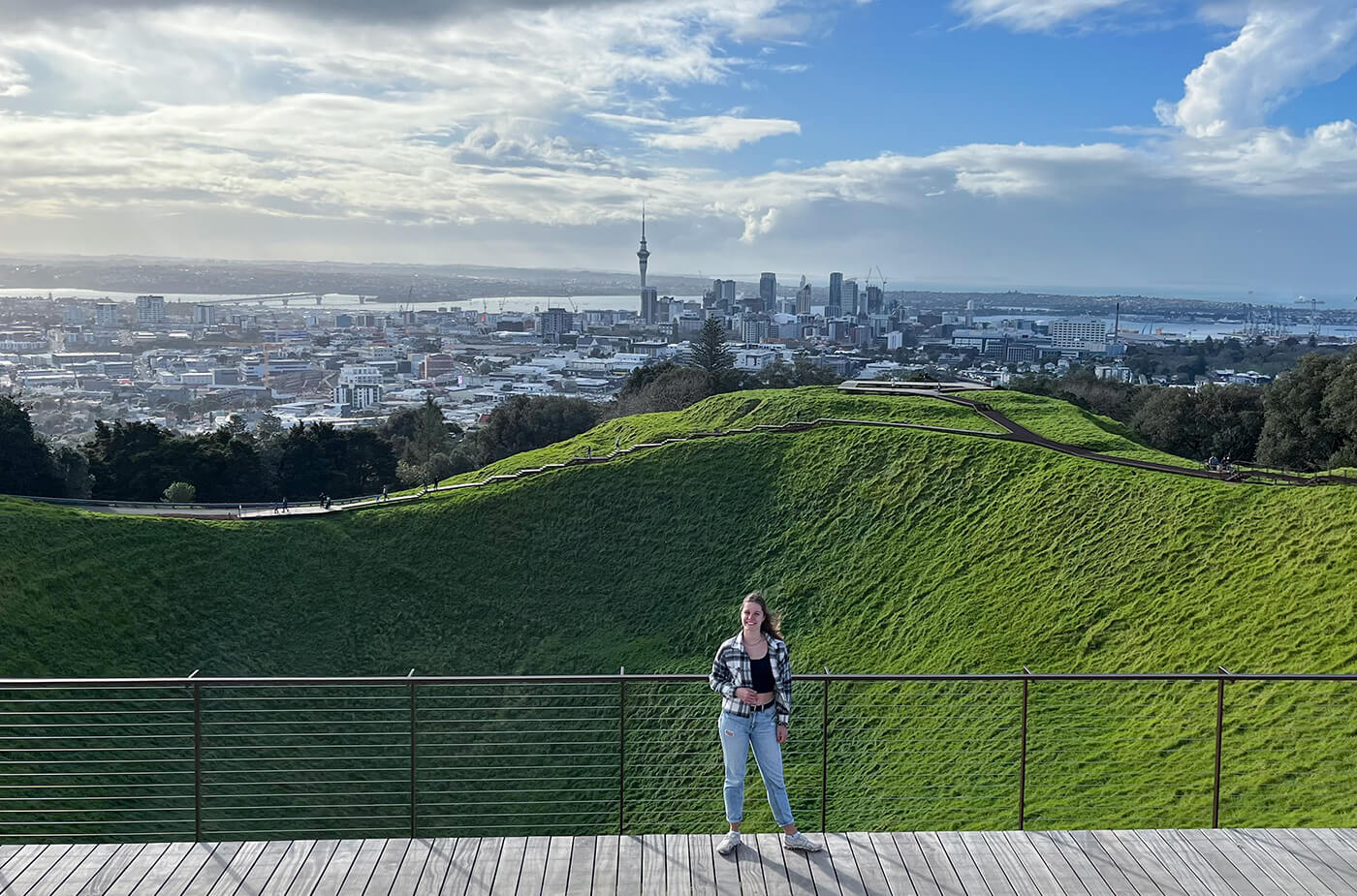 Junge Frau steht auf einem Hügel mit Gras, im Hintergrund sieht man die Skyline von Auckland