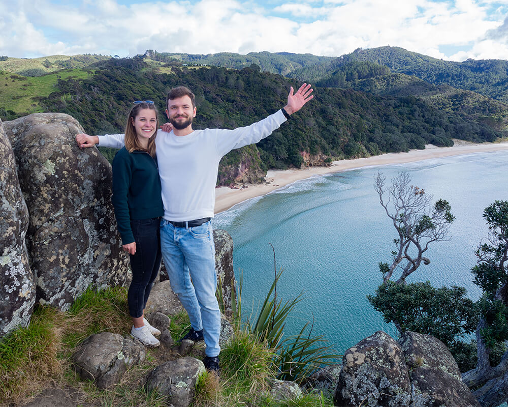 junges Paar steht vor einem Felsen, im Hintergrund sieht man einen Strand mit blauem Meer