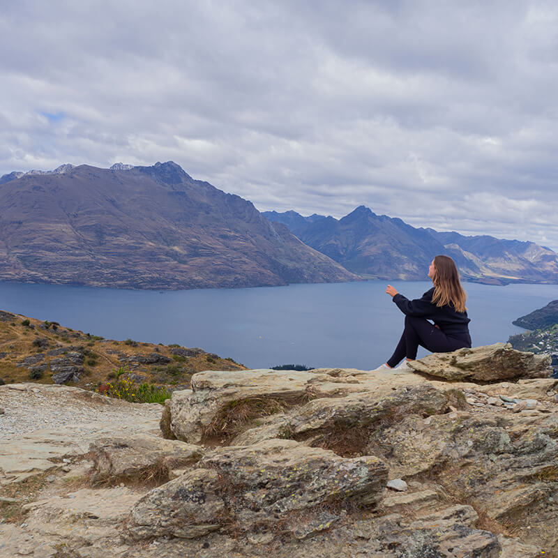 Junge Frau sitzt auf einem Felsen und schaut in die Ferne auf Berge und einen See