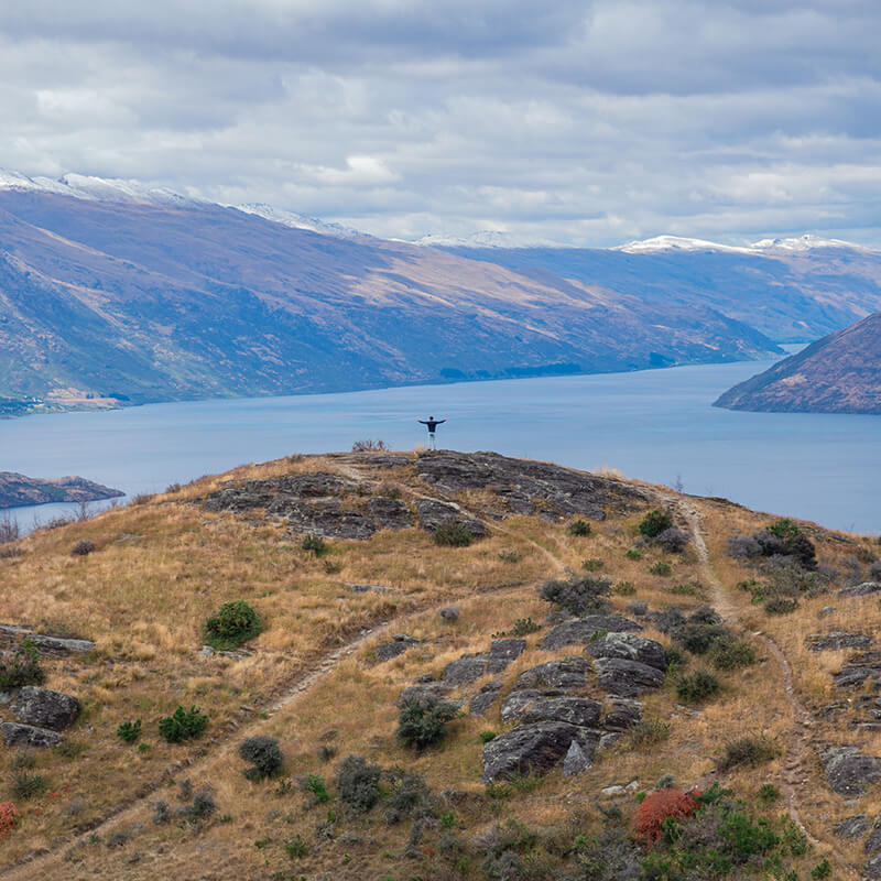 junger Mann steht auf einem Hügel in der Ferne, im Hintergrund der Lake Wakatipu und umliegende Berge
